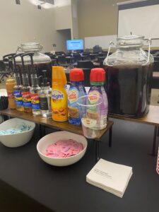 Iced coffe bar with creamers and coffee with ice in a large glass container sitting on a black table