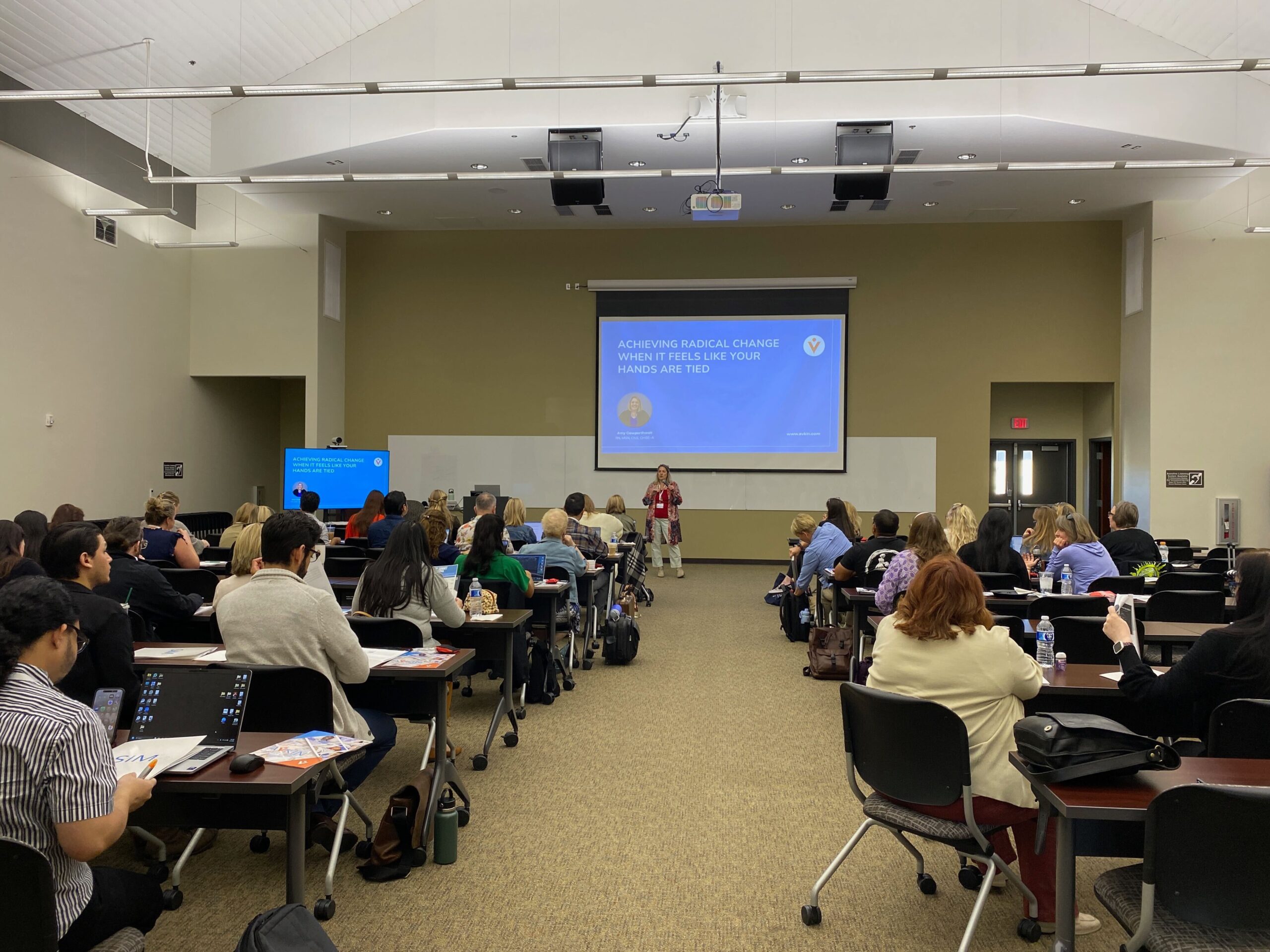 Presenter standing in front of a drop down screen talking to simulationists sitting at long tables.